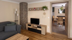 Un salon comprenant un canapé, une table basse, un téléviseur posé sur un meuble en bois, une étagère murale ornée de plantes et un escalier menant à une salle à manger.