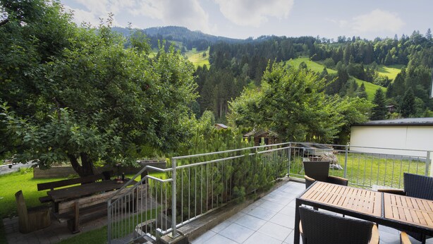 Une terrasse avec une table et des chaises en bois, une rambarde en métal et une vue sur un paysage verdoyant, avec des arbres et des collines.