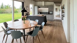 Eine moderne Küche mit einer zentralen Kochinsel, einem Essbereich mit Holztisch und -stühlen sowie Blick auf einen grünen Garten durch große Fenster.