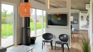 Ein modernes Wohnzimmer mit Kamin, zwei Sesseln, einem Fernseher und einem großen Fenster mit Blick auf einen Garten.
