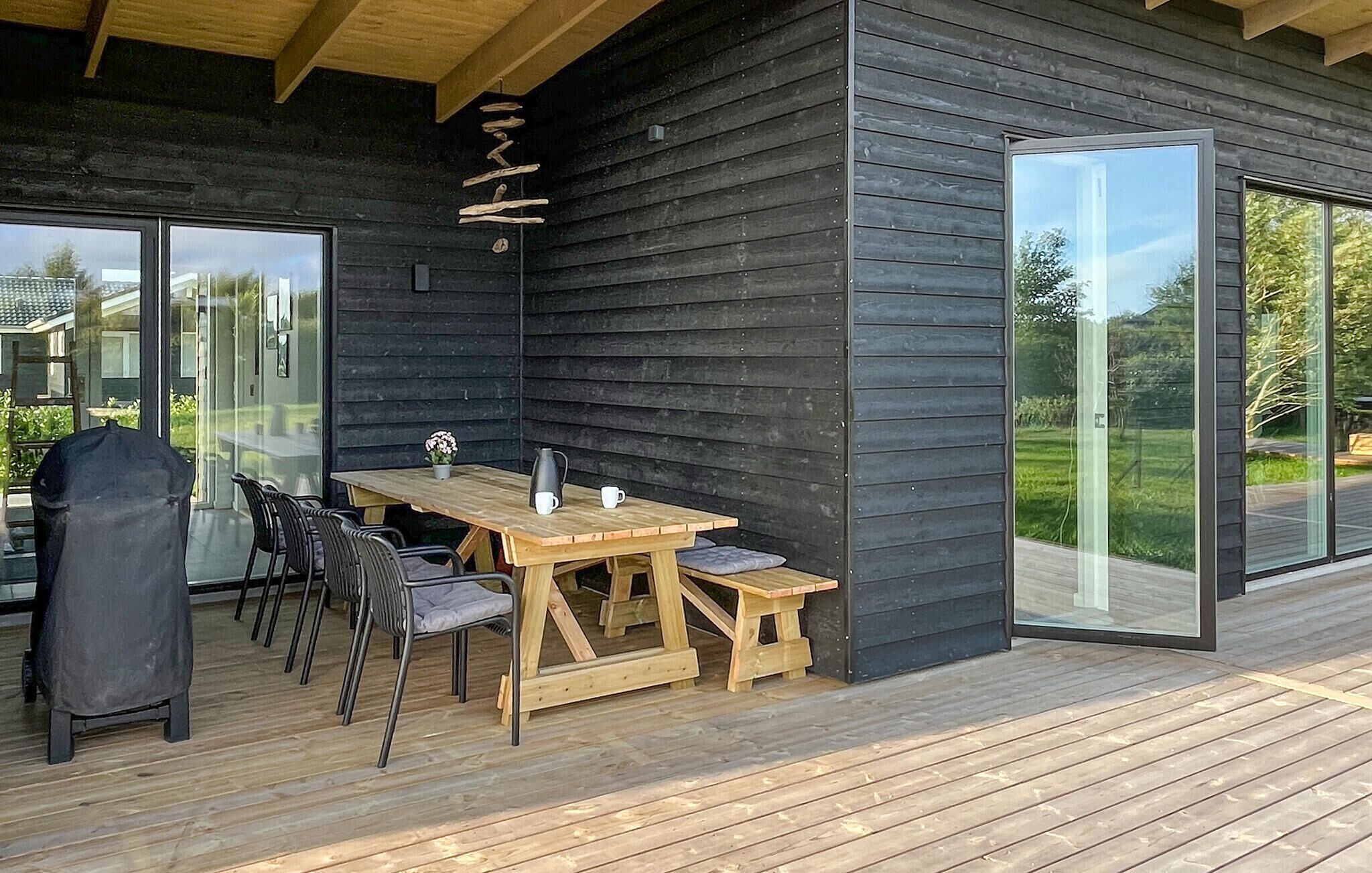 Una terraza de madera al aire libre con una mesa, sillas, una parrilla y un ventanal de vidrio que da a un jardín.