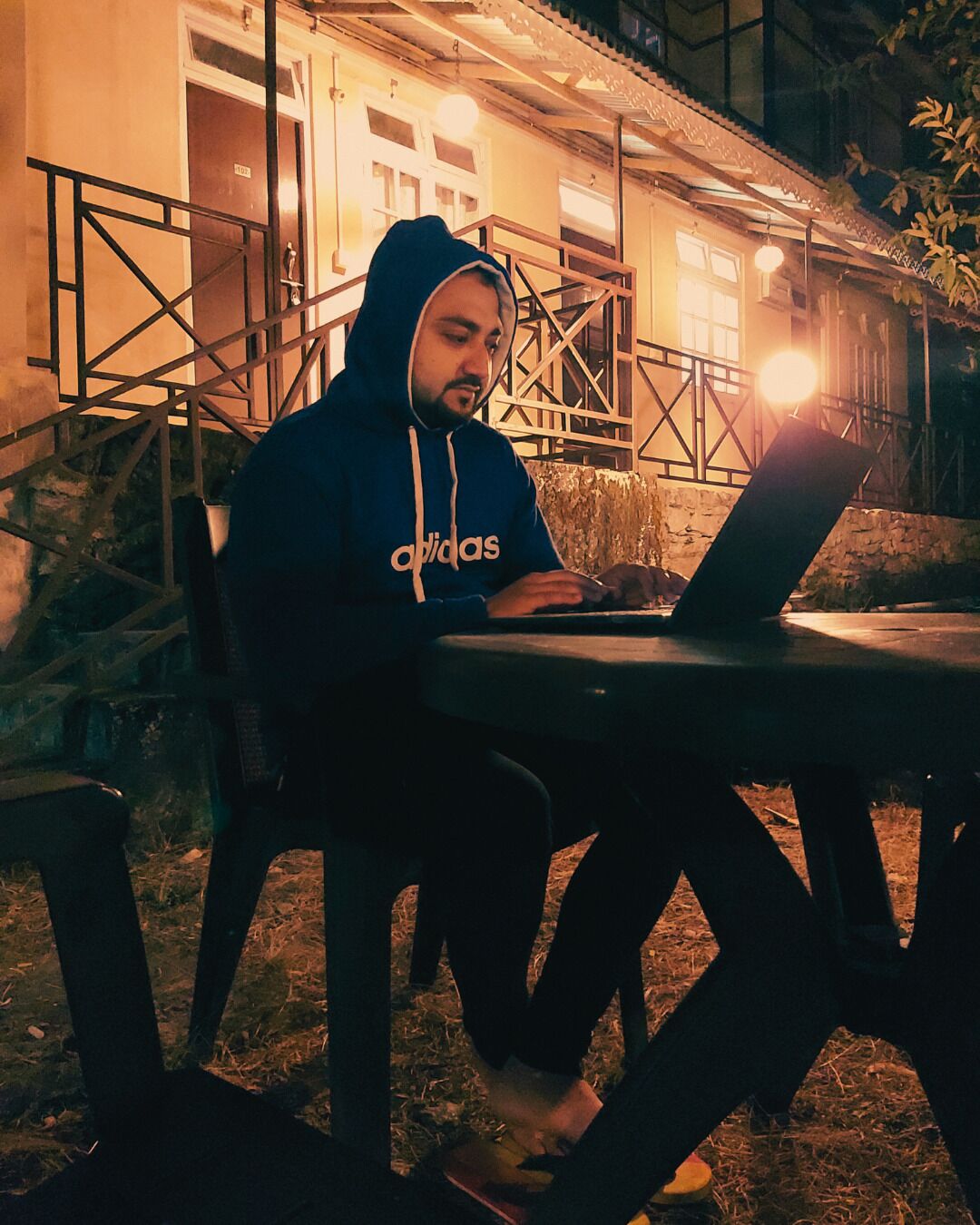A person sitting at an outdoor table with a laptop, wearing a hoodie, in front of a building with a staircase and a railing.