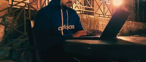 A person sitting at an outdoor table with a laptop, wearing a hoodie, in front of a building with a staircase and a railing.