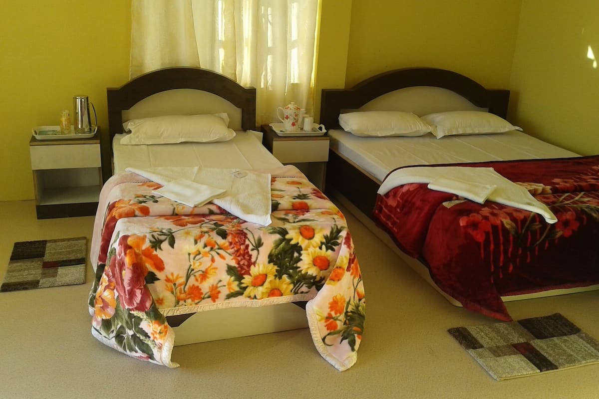 A hotel room with two single beds, each with a floral bedspread and white pillows. There are bedside tables with lamps and a small table with a pitcher and cups. The room has a carpeted floor and a window with curtains.