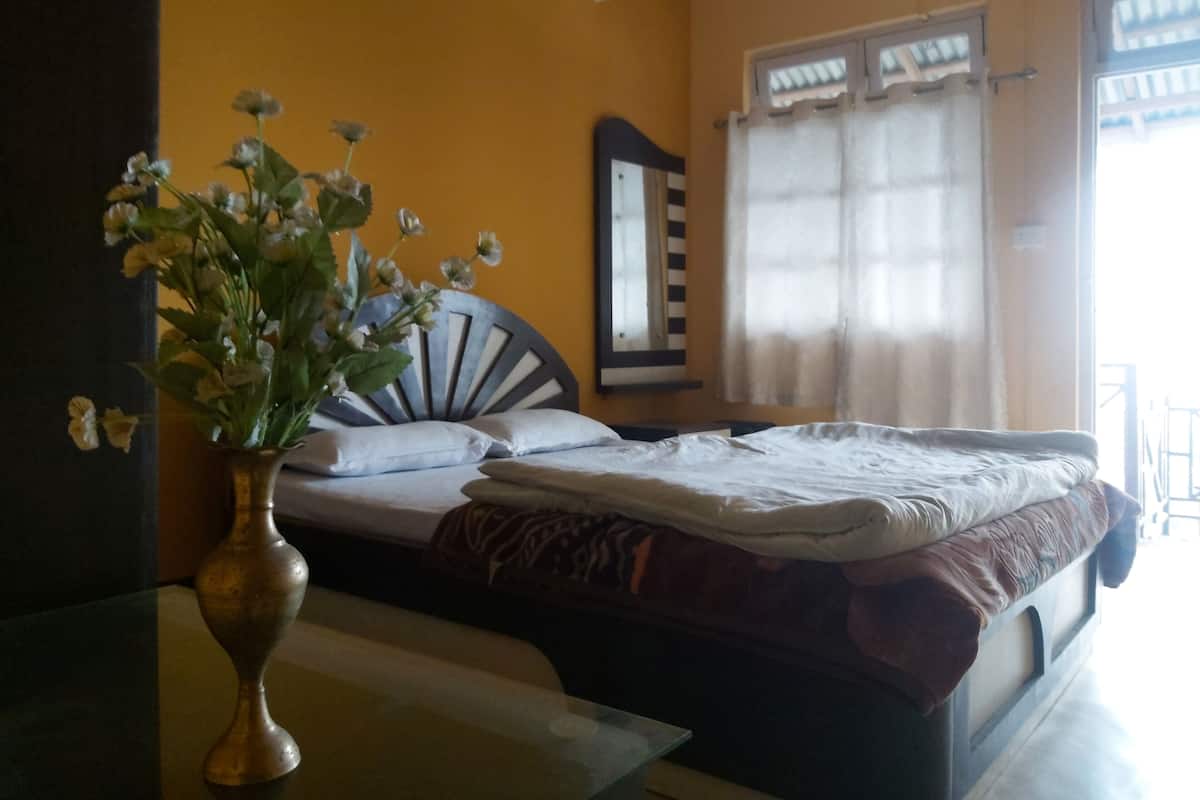 A bedroom with a bed, a bedside table, a vase with flowers, a mirror, and a window with curtains.