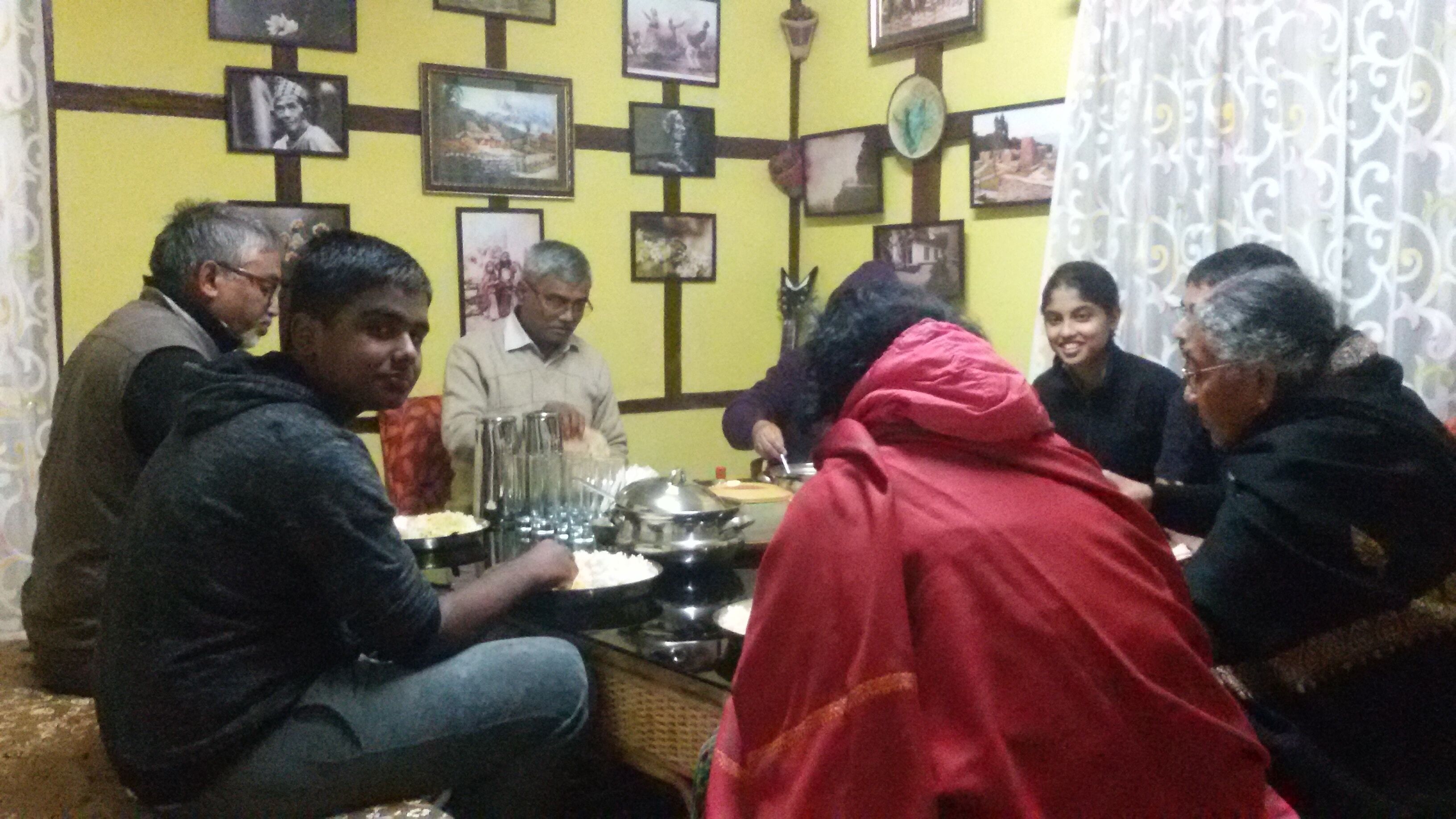 A group of people seated around a table with a traditional meal setup, including a pot and glasses, in a room with a yellow wall and framed pictures.