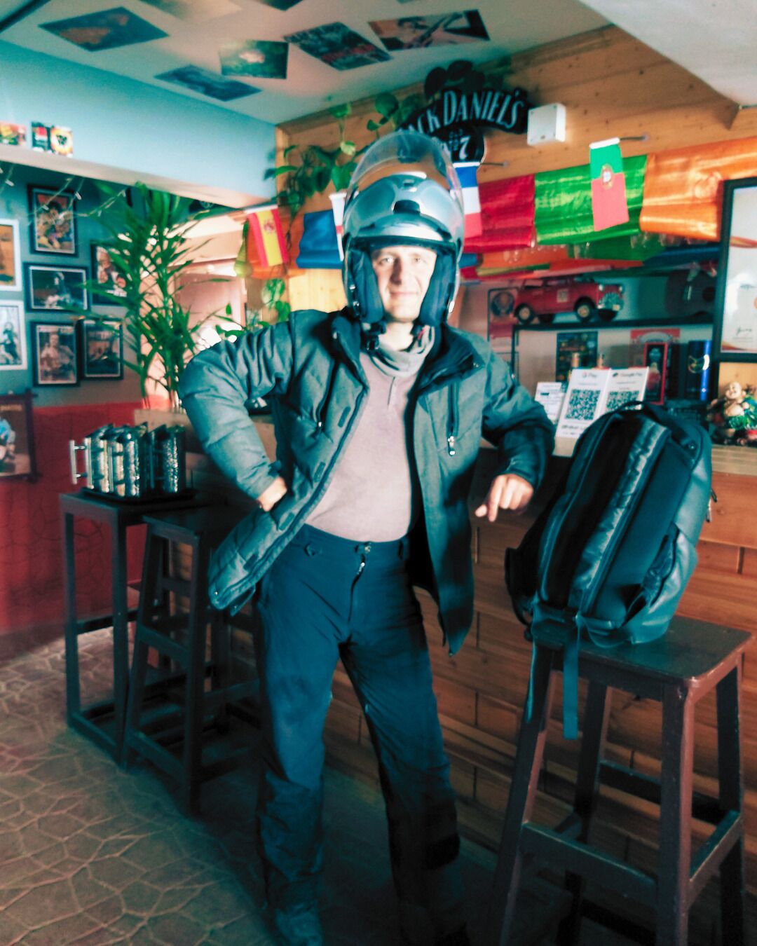 A person in a motorcycle helmet and jacket standing in a room with a wooden floor, a table with a backpack, and various items on shelves in the background.