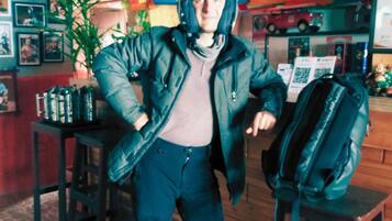 A person in a motorcycle helmet and jacket standing in a room with a wooden floor, a table with a backpack, and various items on shelves in the background.