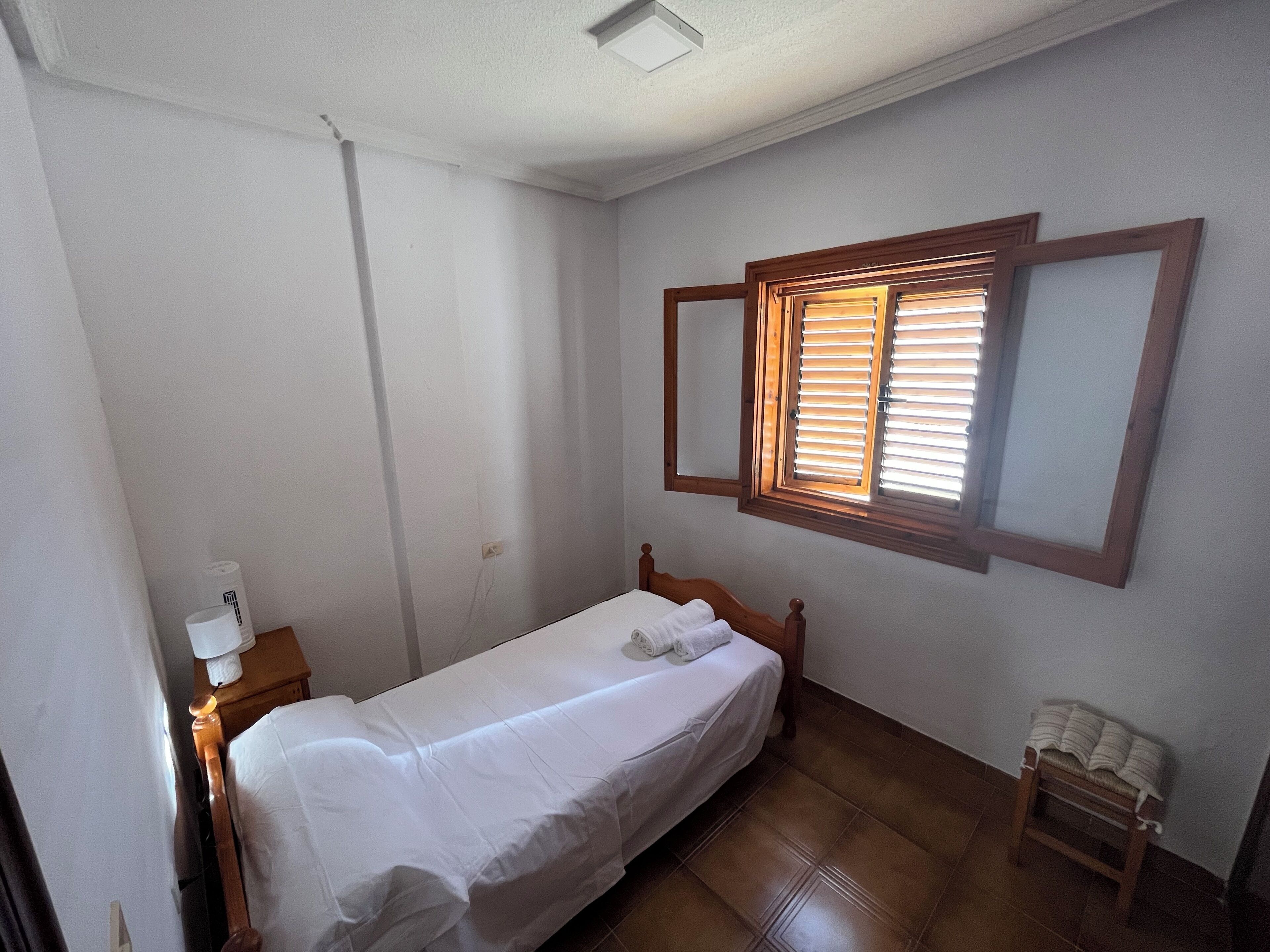 A small, single-bed room with a wooden headboard, a bedside table with a lamp, and a window with blinds.