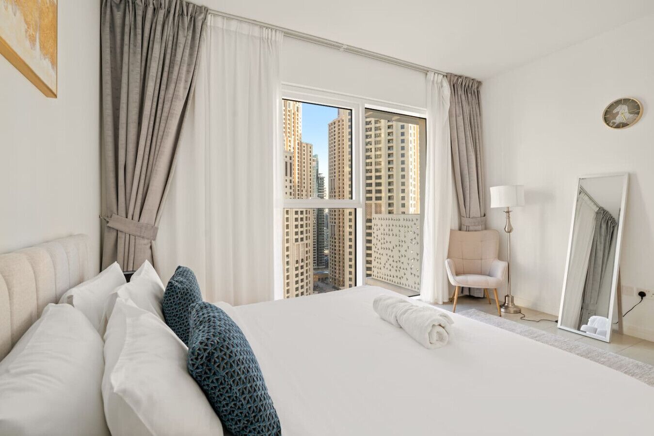 Habitación de hotel con una cama grande, una silla, una lámpara, un espejo y vistas a edificios altos a través de la ventana.