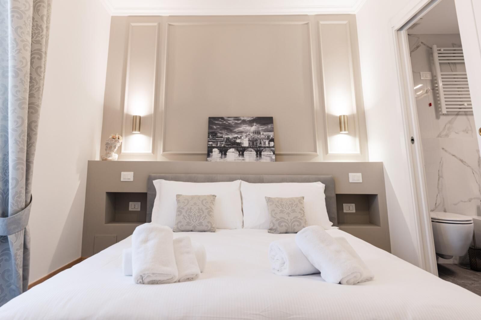 Un lit bien fait, avec du linge de lit blanc et des serviettes pliées, une tête de lit ornée d’un tableau représentant un paysage urbain, et deux lampes fixées au mur.
