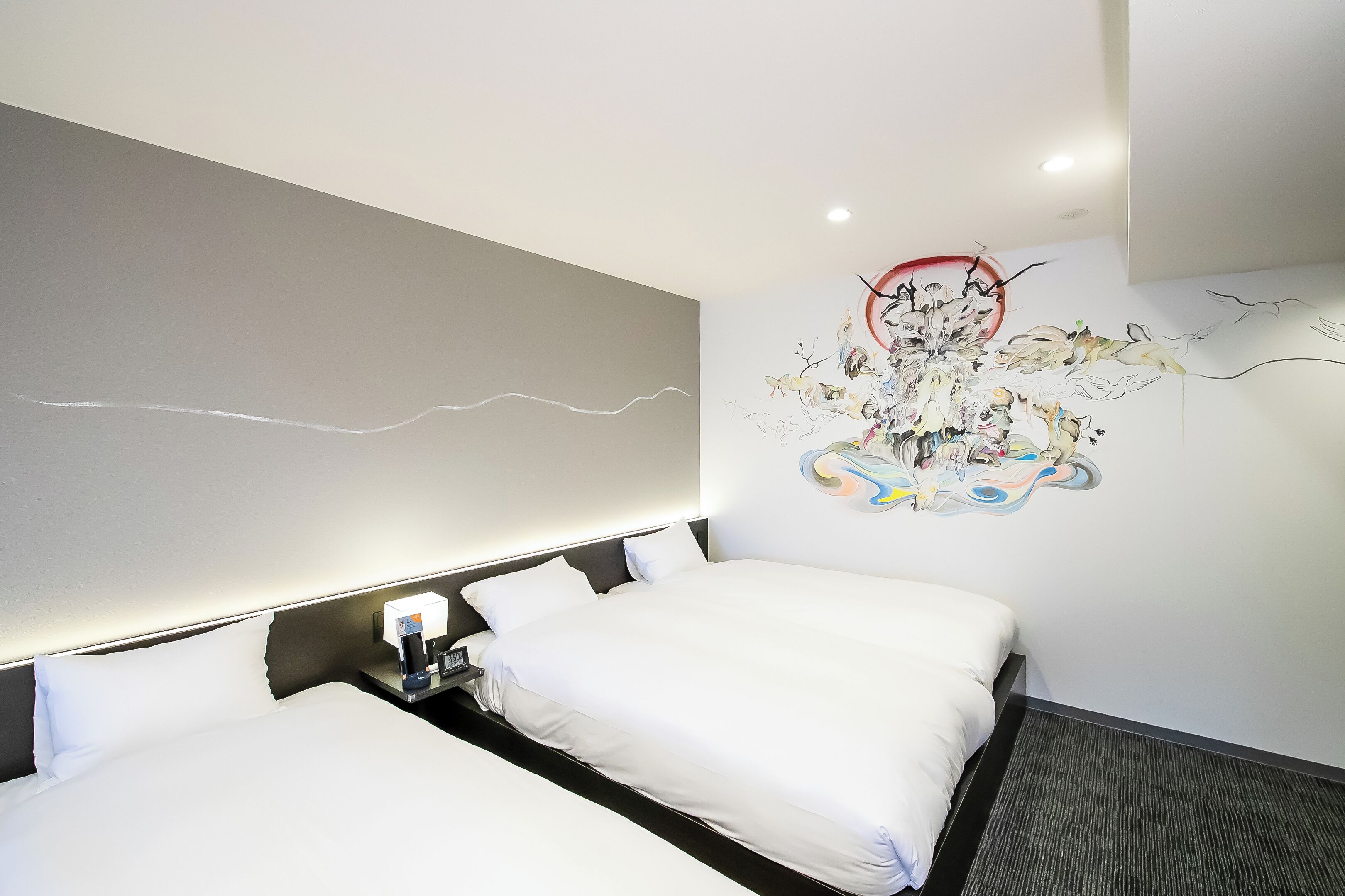 Une chambre d’hôtel moderne avec un grand lit, une table de chevet avec une lampe et un mur décoré d’œuvres d’art abstraites.
