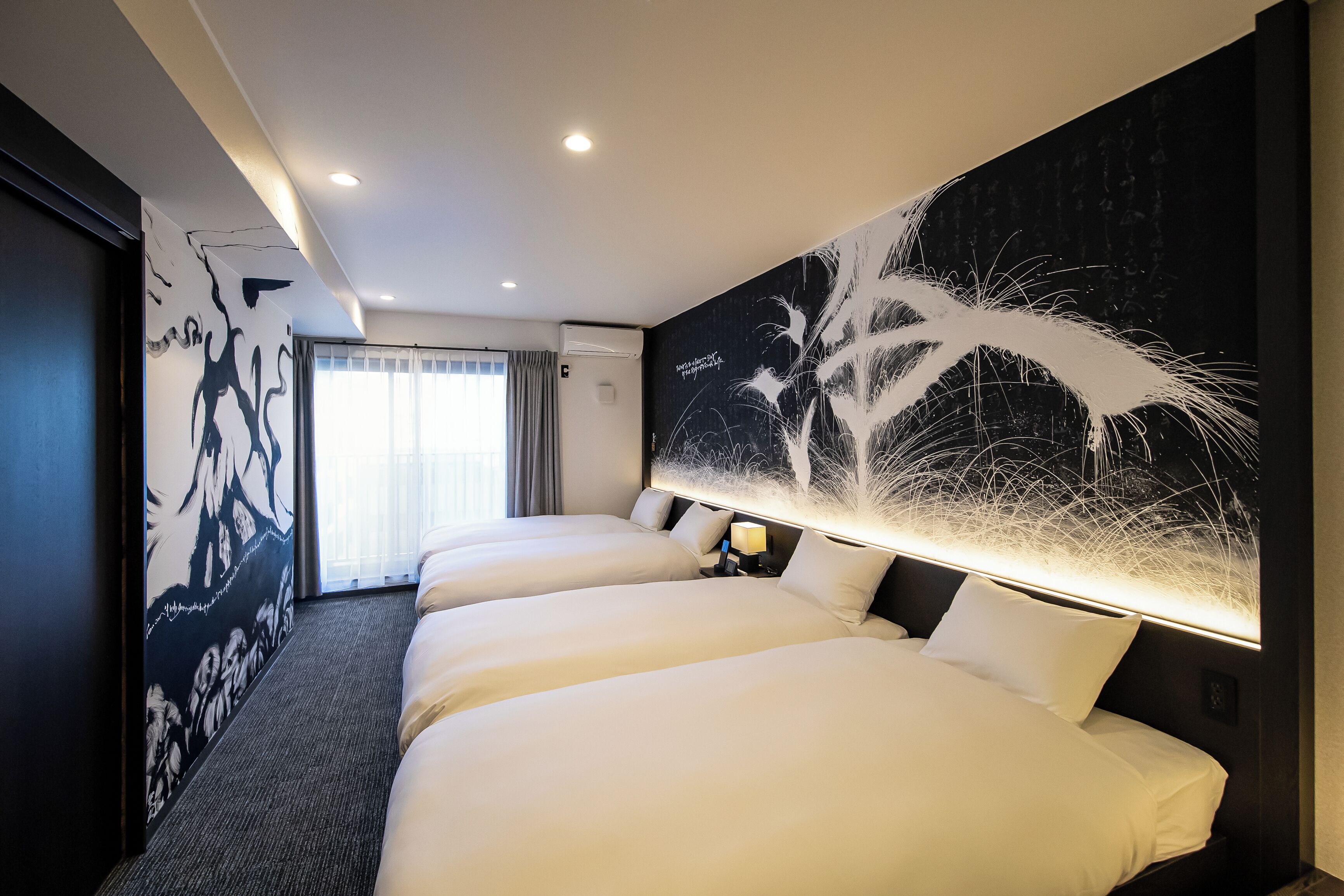 Una habitación de hotel con tres camas, un cuadro abstracto grande en la pared y vistas al exterior a través de una ventana con cortinas.