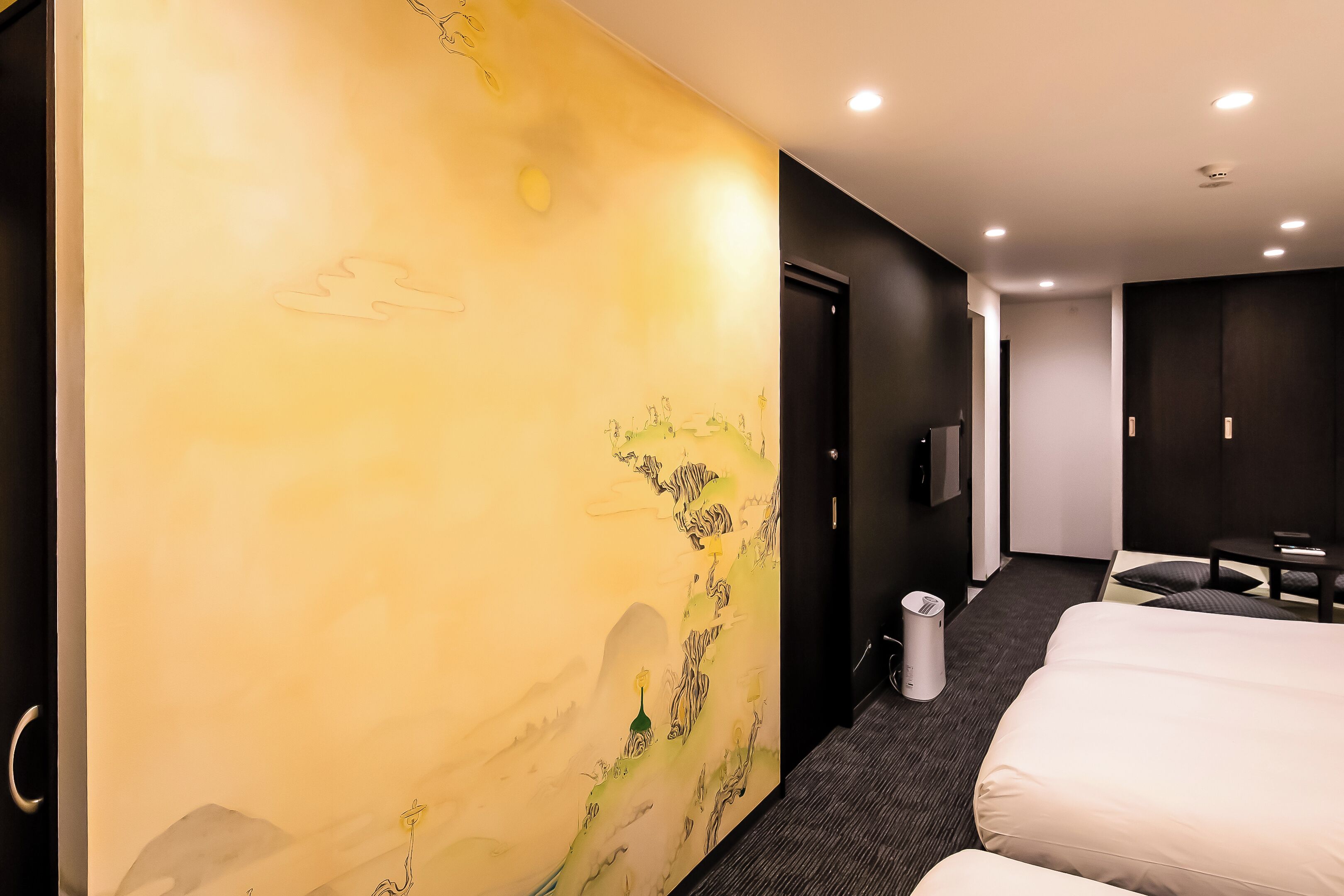 A hotel room with a large mural on the wall, a bed, a desk, and a trash bin.