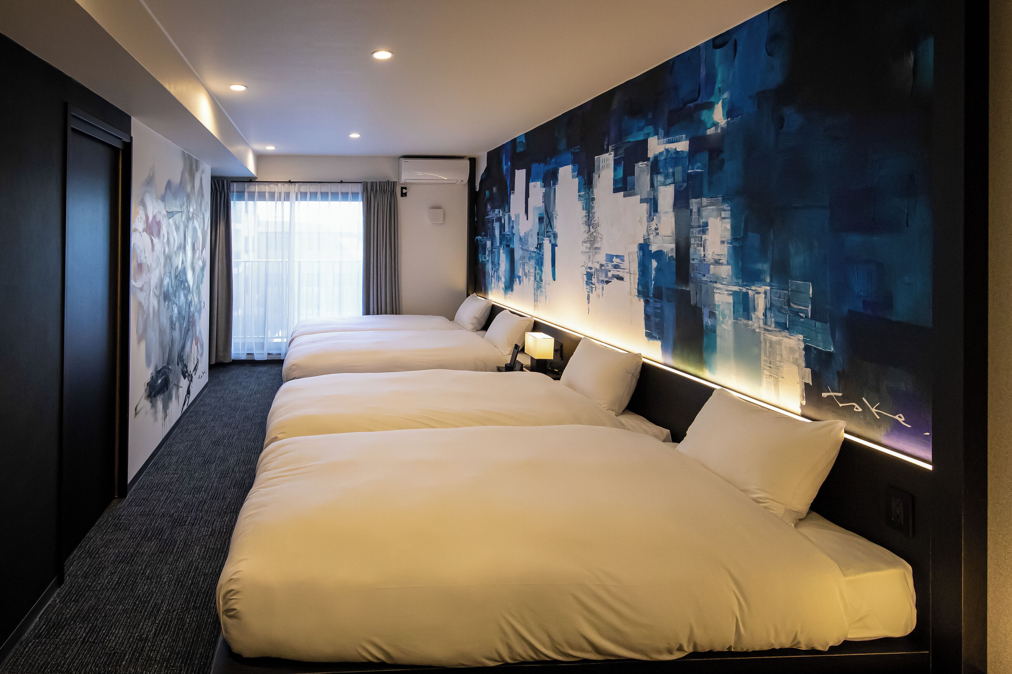 Une chambre d’hôtel avec trois lits, une grande peinture abstraite au mur et une vue sur le paysage urbain par la fenêtre.