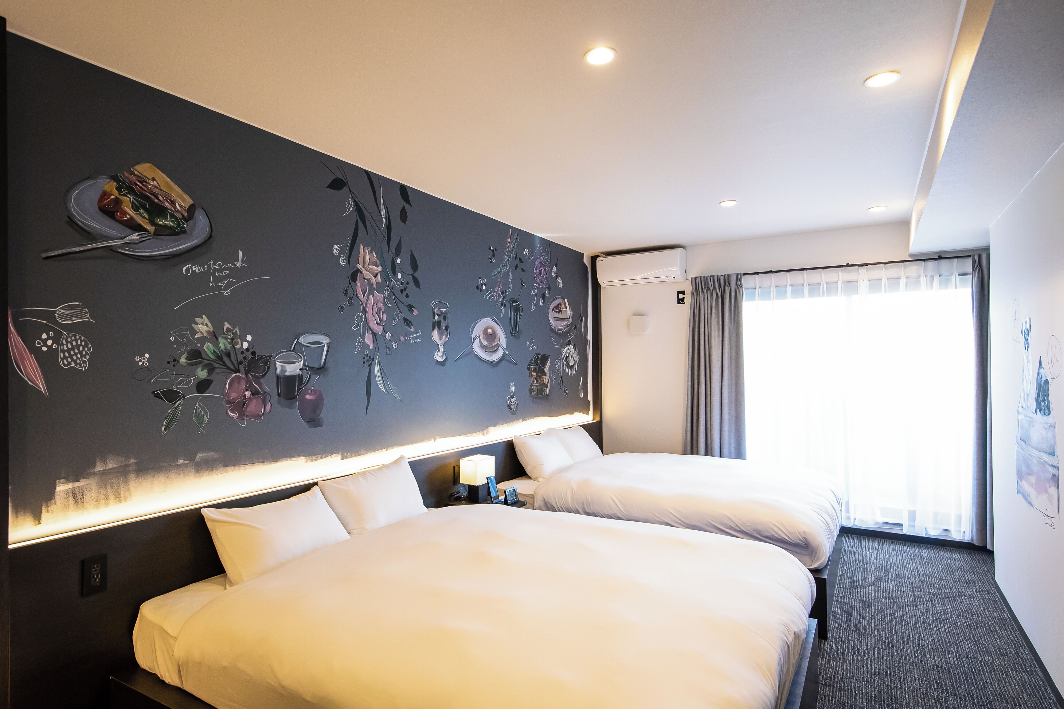 A hotel room with two beds, a dark wall with floral and food illustrations, a nightstand with a lamp, and a window with curtains.