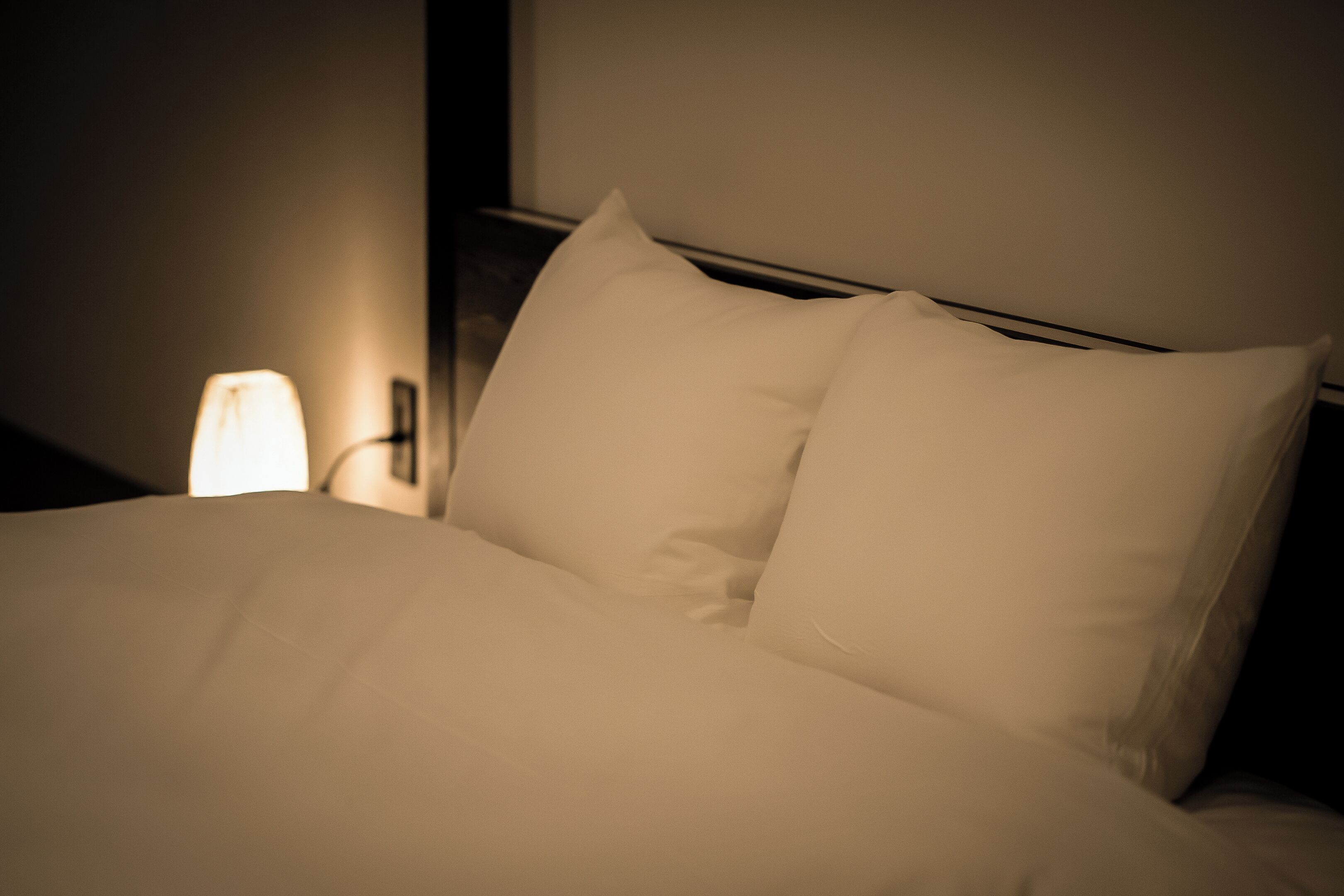 Una cama bien tendida con sábanas y almohadas blancas, una lámpara de noche y una lámpara de pared.
