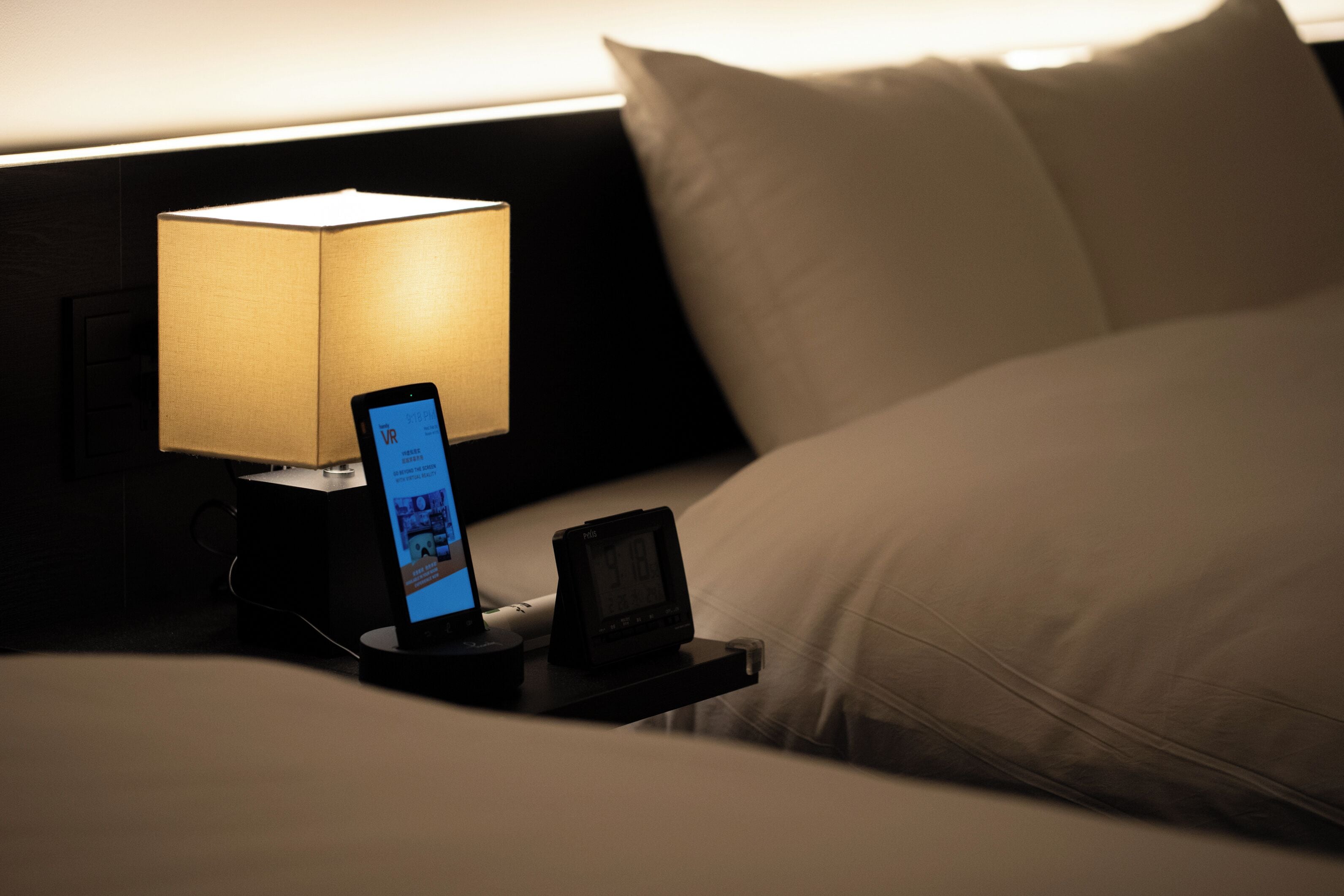 Un lit bien fait, avec des draps et des oreillers blancs, une table de chevet avec une lampe et un smartphone affichant une application de réalité virtuelle.
