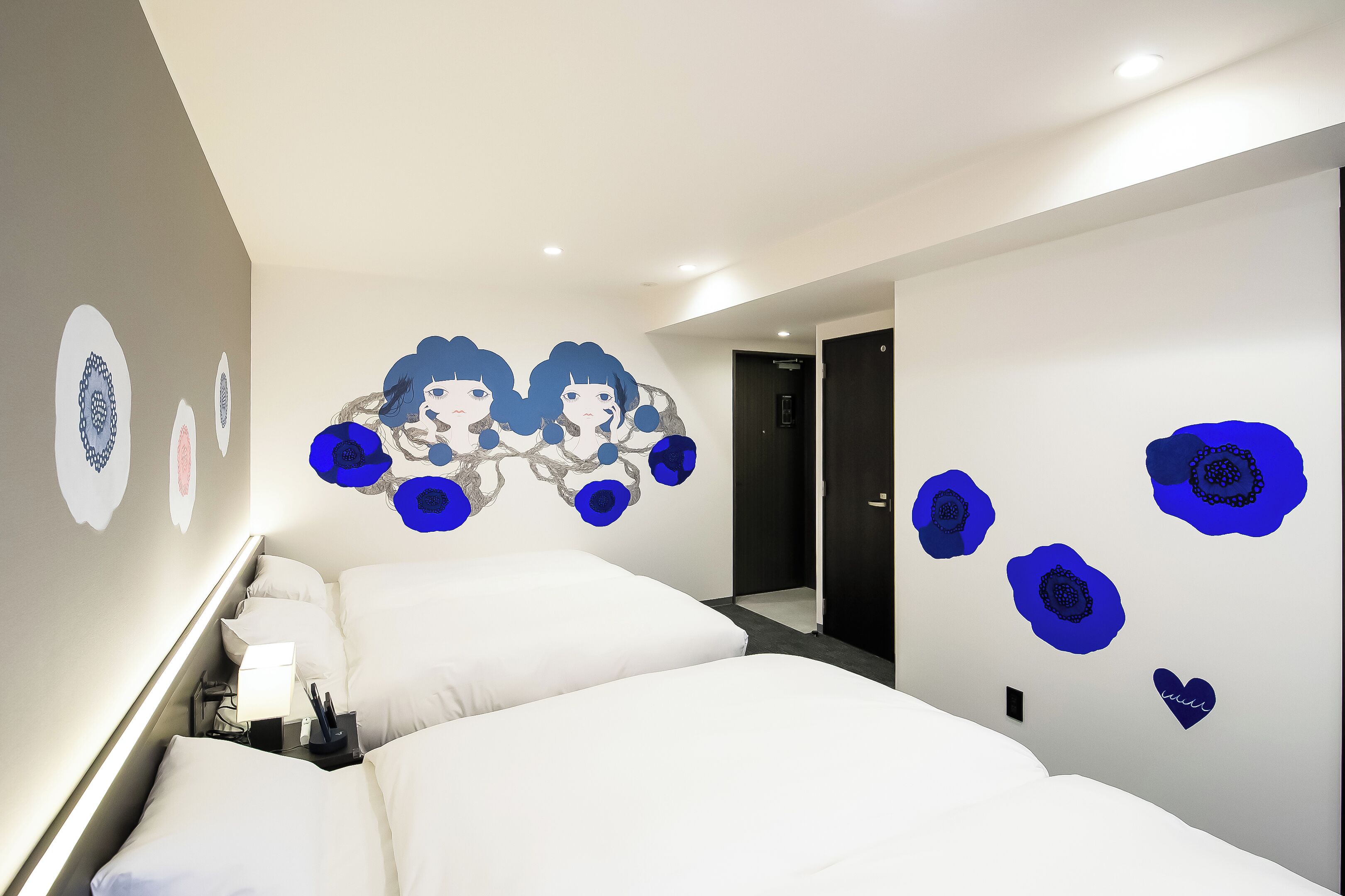 A hotel room with two beds, a nightstand, a wall with decorative blue floral decals, and a door to the hallway.