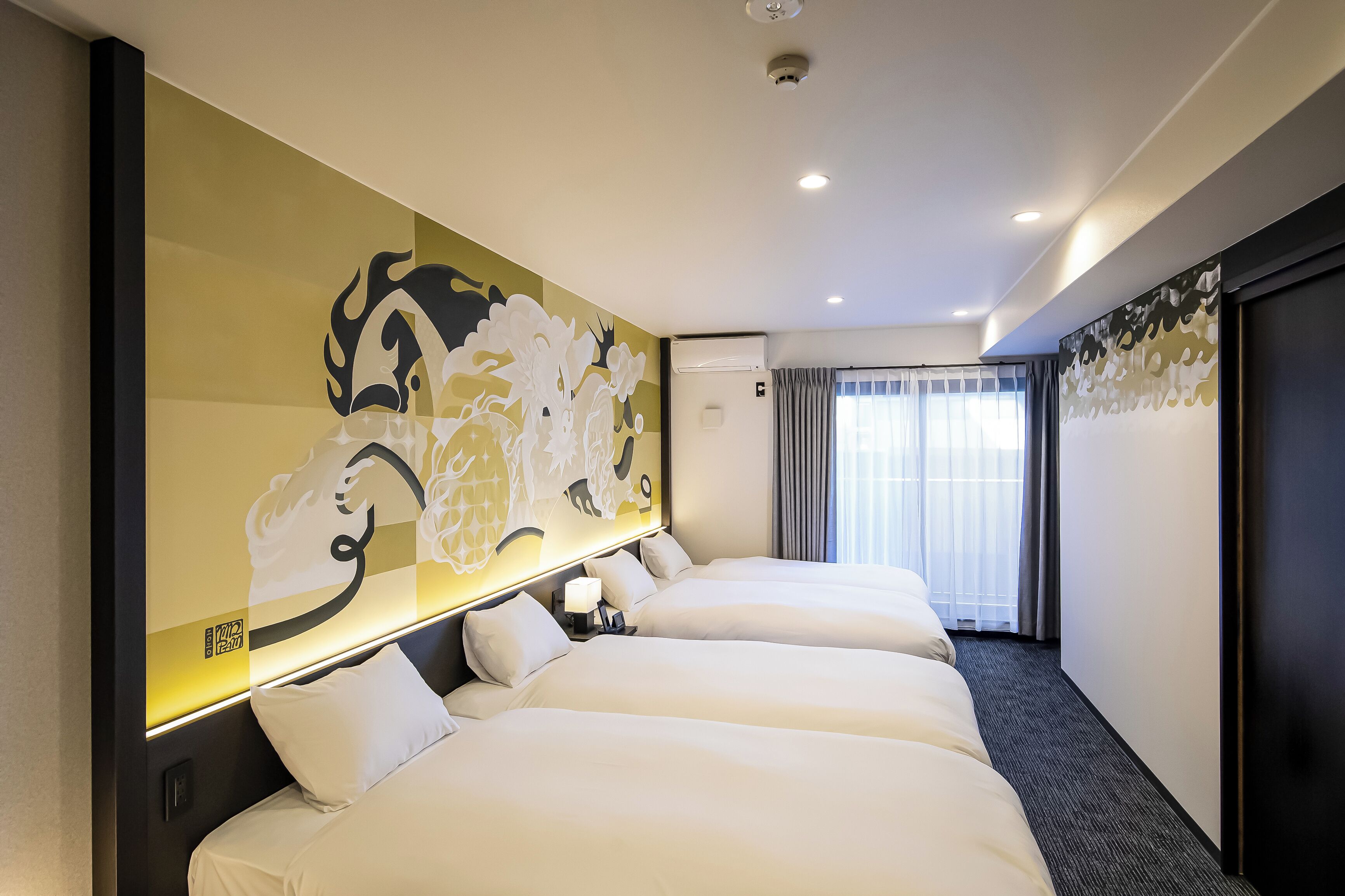 A hotel room with three beds, a large abstract mural on the wall, a modern air conditioning unit, and a view of the outdoors through a window with curtains.