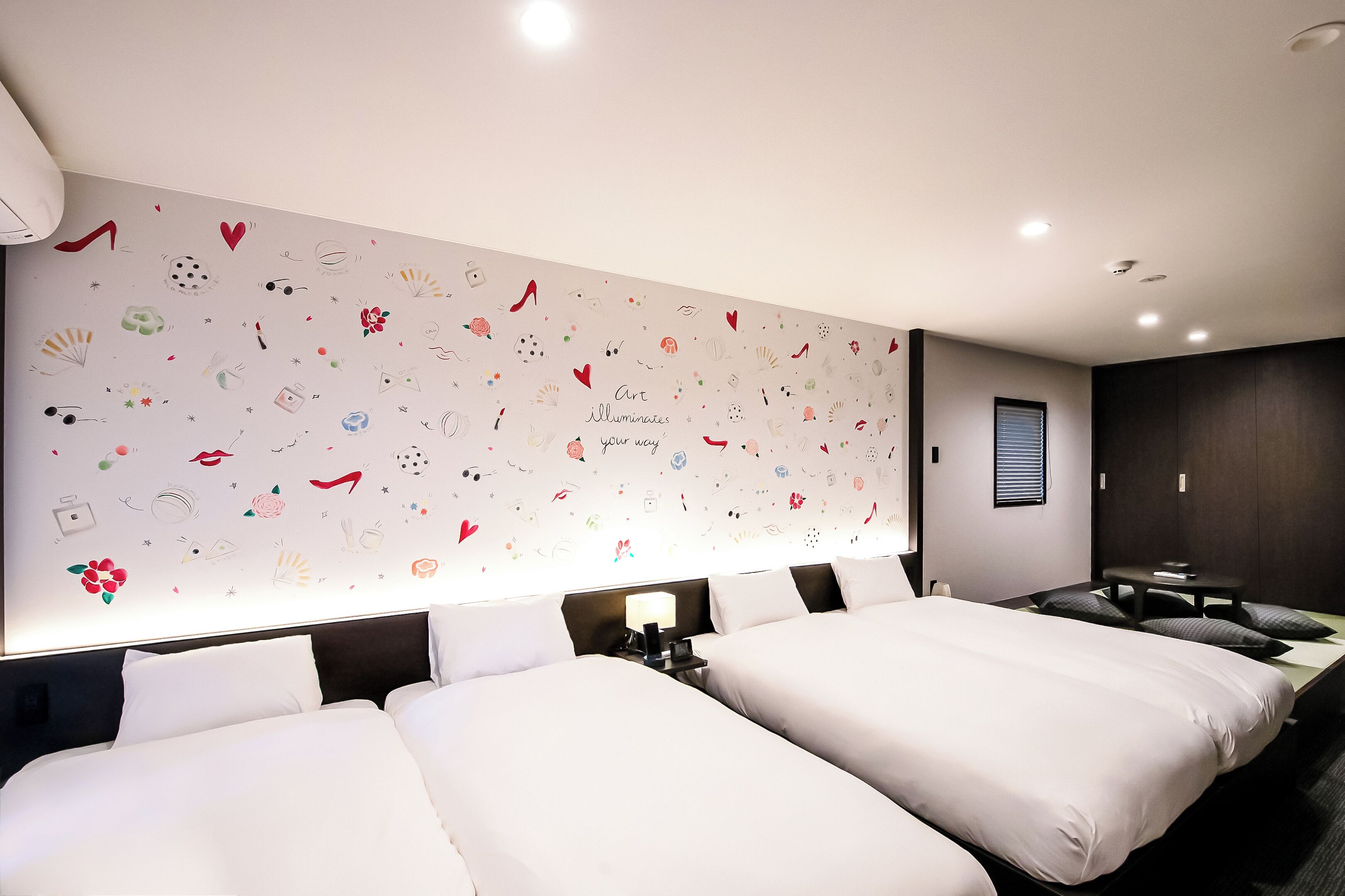 Quarto de hotel com duas camas, um grande mural na parede, uma escrivaninha e uma janela.