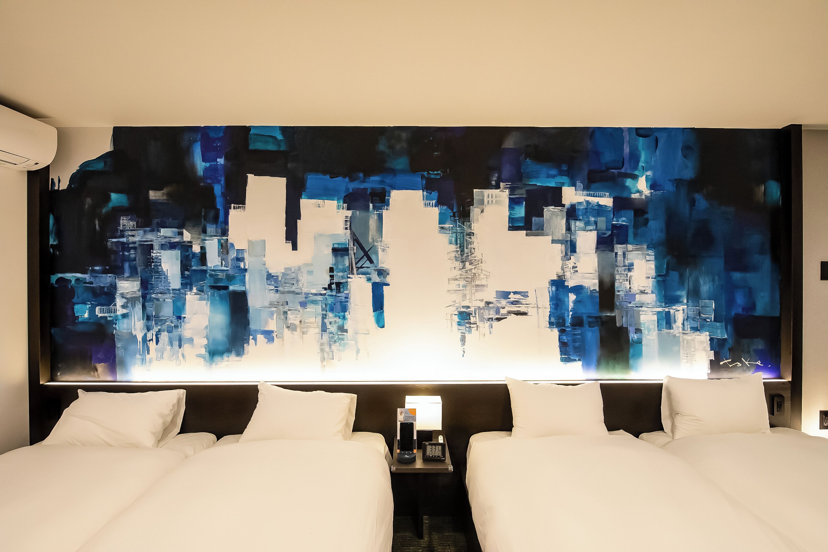 A hotel room with two beds, a large abstract painting on the wall, and a nightstand with a lamp and phone.