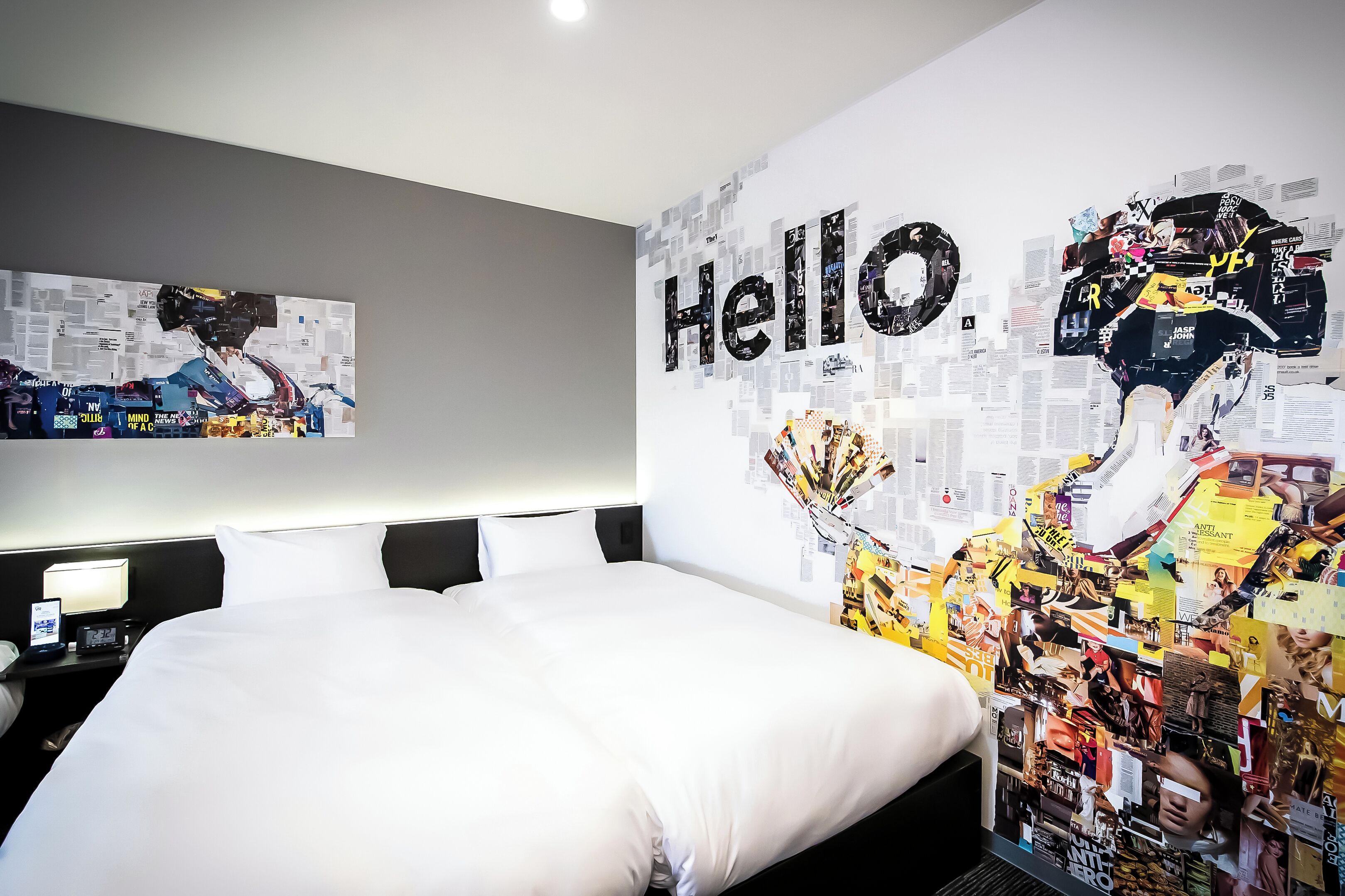 Ein Hotelzimmer mit einem großen Bett, einem Nachttisch mit Lampe und einer Wand mit einer Collage aus Bildern und dem Wort 'Hello'.