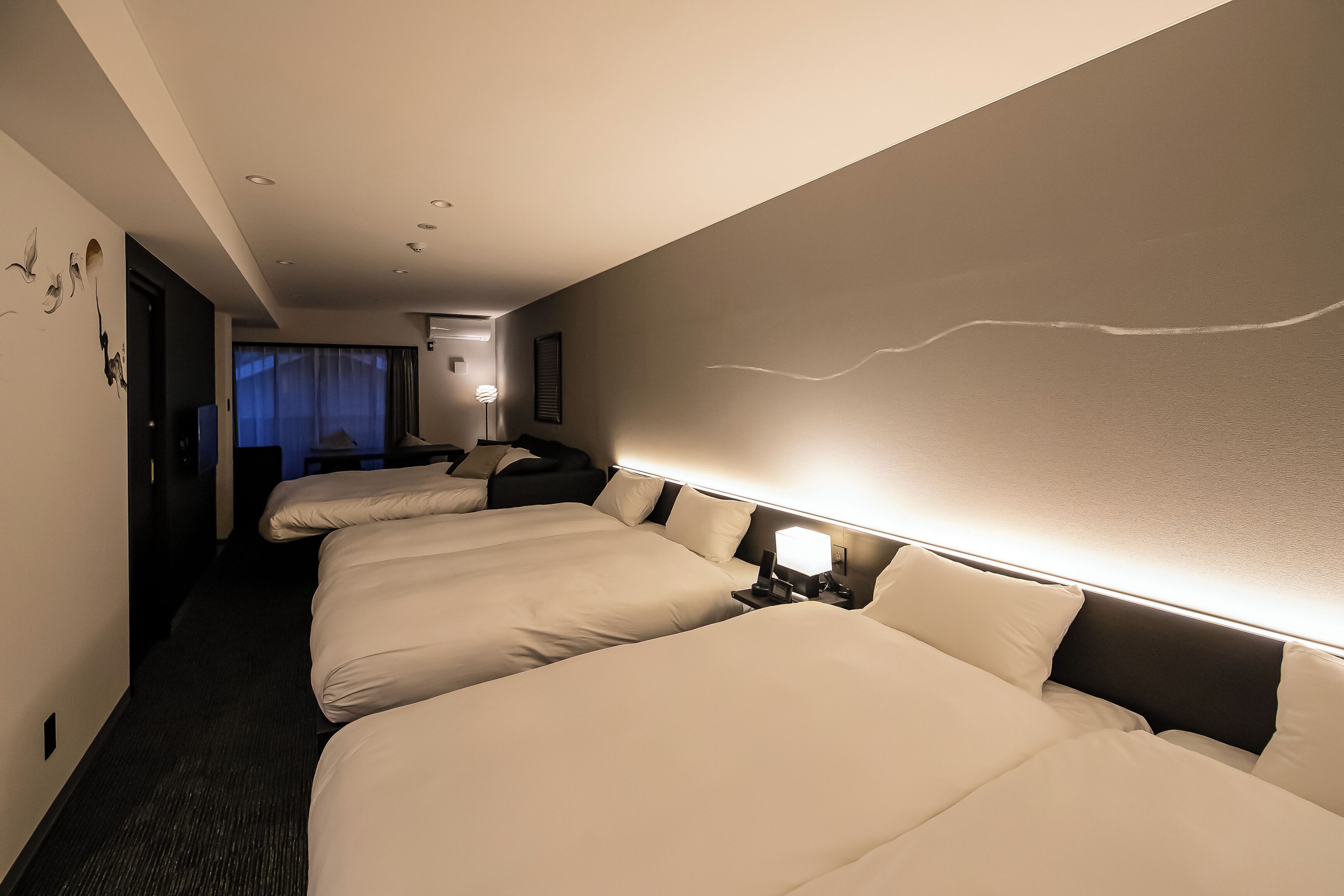 Une chambre d’hôtel avec trois lits, une table de chevet avec une lampe et une vue sur la ville la nuit.