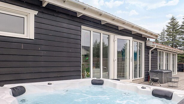 A hot tub in a backyard with a dark wooden house, sliding glass doors, and a patio area with outdoor furniture. - 4 Star Holiday Home in Vaeggerlose (Vaeggerlose)