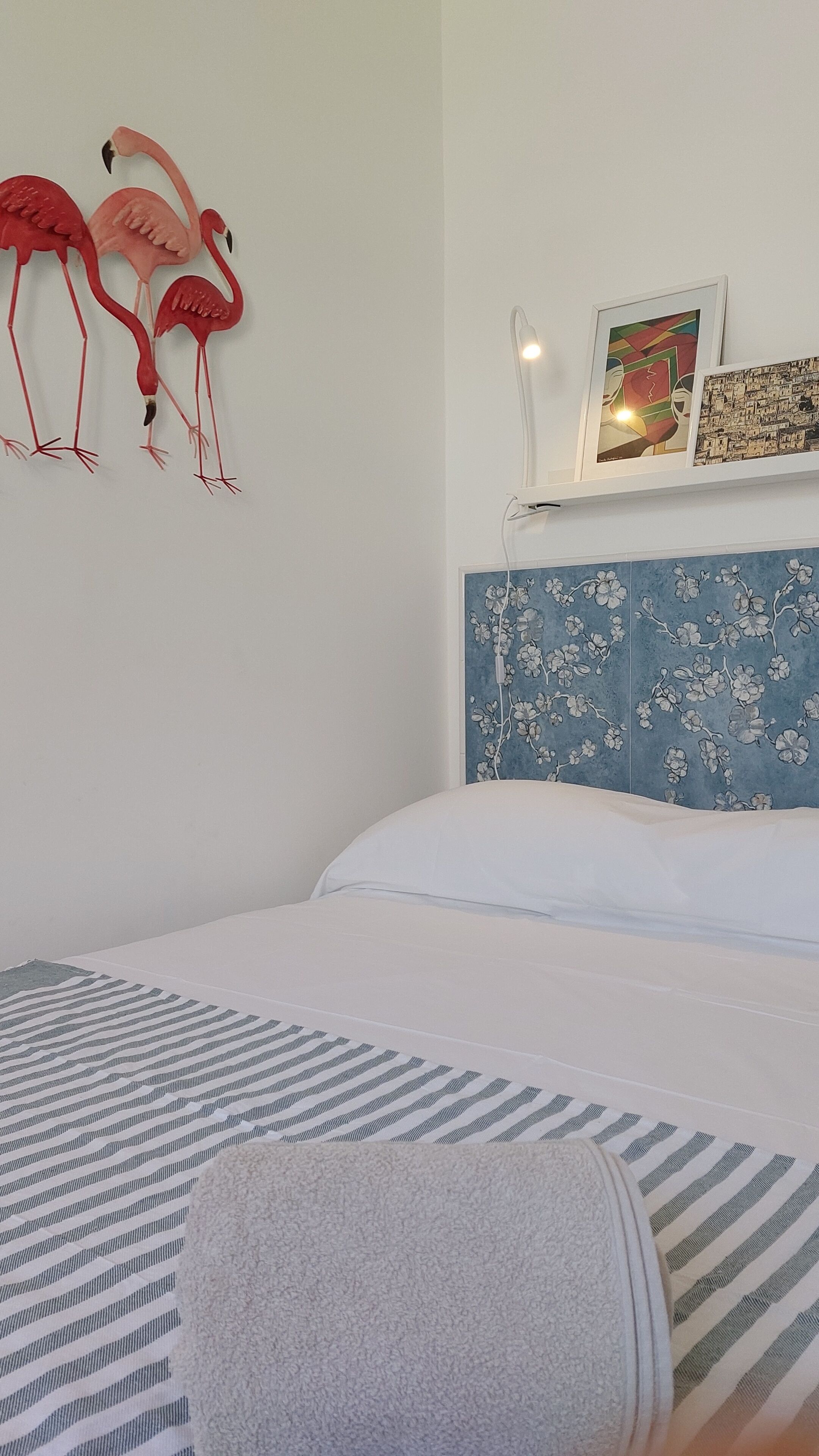 Quarto com cama, prateleira na parede com duas fotos emolduradas e decoração de flamingos.