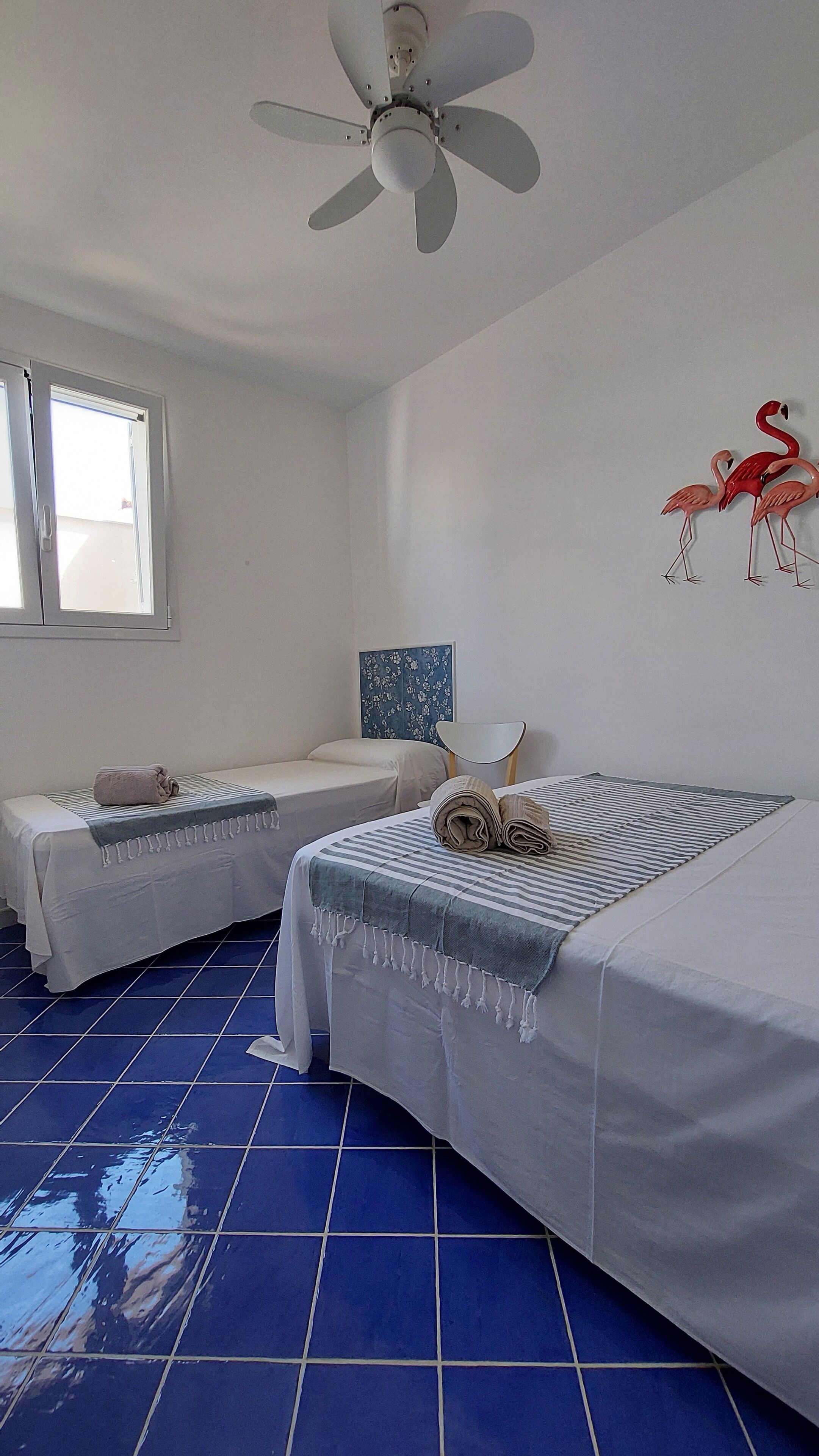 Habitación de hotel con dos camas, ventilador de techo, ventana y un adorno de flamencos en la pared.