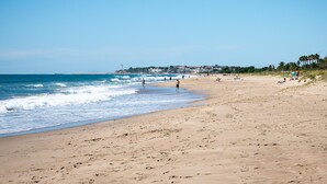 A wide sandy beach with gentle waves, a clear sky, and a distant town with buildings and palm trees.