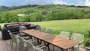 An outdoor patio with a wooden table and white chairs, a wicker sofa, and a view of rolling hills. - Renovated country chic cottage, 6 bedrooms for 13, garden, terrace, Haute-Loire (Siaugues-Sainte-Marie)