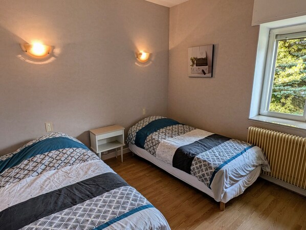 A bedroom with two beds, a small bedside table, a radiator, and a window with a view of greenery.