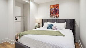 Un dormitorio ordenado con una cama, un ventilador de techo, un cartel en la pared y una mesita de noche con una lámpara.