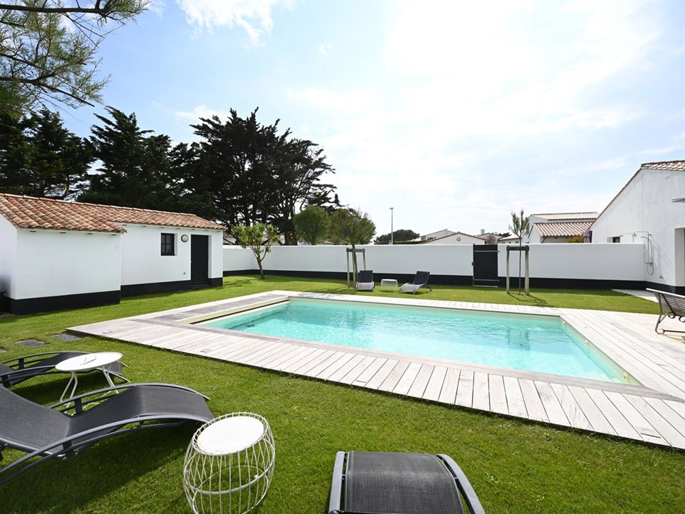 Une piscine rectangulaire avec une terrasse en bois, entourée d’une clôture blanche, et aménagée avec des chaises longues et une petite table.