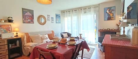 Een eetgedeelte met een tafel voor vier personen, een bank, een kitchenette en een balkon met gordijnen.
