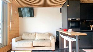 Un espace de vie moderne avec un plafond en bois, un canapé, un coin cuisine équipé d’un téléviseur et une table à manger agrémentée de bols rouges.