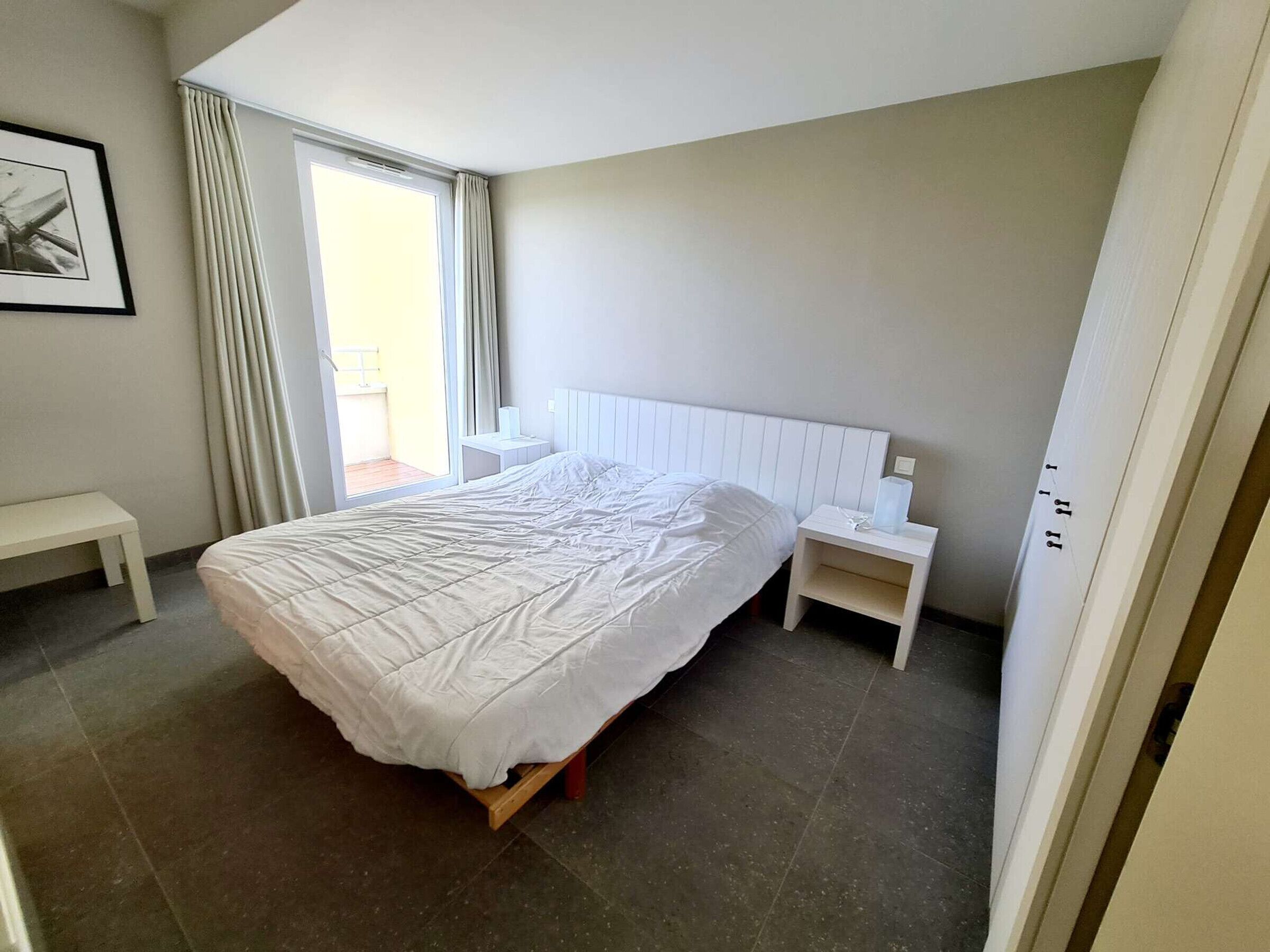 Een slaapkamer met een groot bed, een nachtkastje, een bankje en een raam met gordijnen.