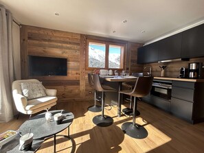 A compact kitchen with wooden walls, a dining area with modern chairs, a flat-screen TV, and a window with a mountain view.