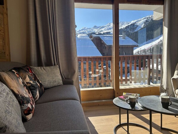 A cozy room with a gray sofa, a small round table with two cups, and a view of snow-covered mountains and a lake.
