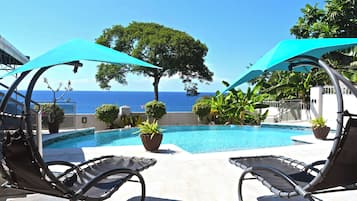 Un espace aménagé au bord de la piscine, avec deux chaises longues, un grand arbre et une vue sur l’océan.