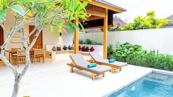 A poolside area with two sun loungers, a wooden pergola, a dining area with a table and chairs, and a small garden with plants.