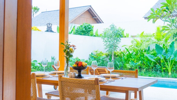 A dining area with a poolside view, featuring a table set with plates, glasses, and a fruit bowl.