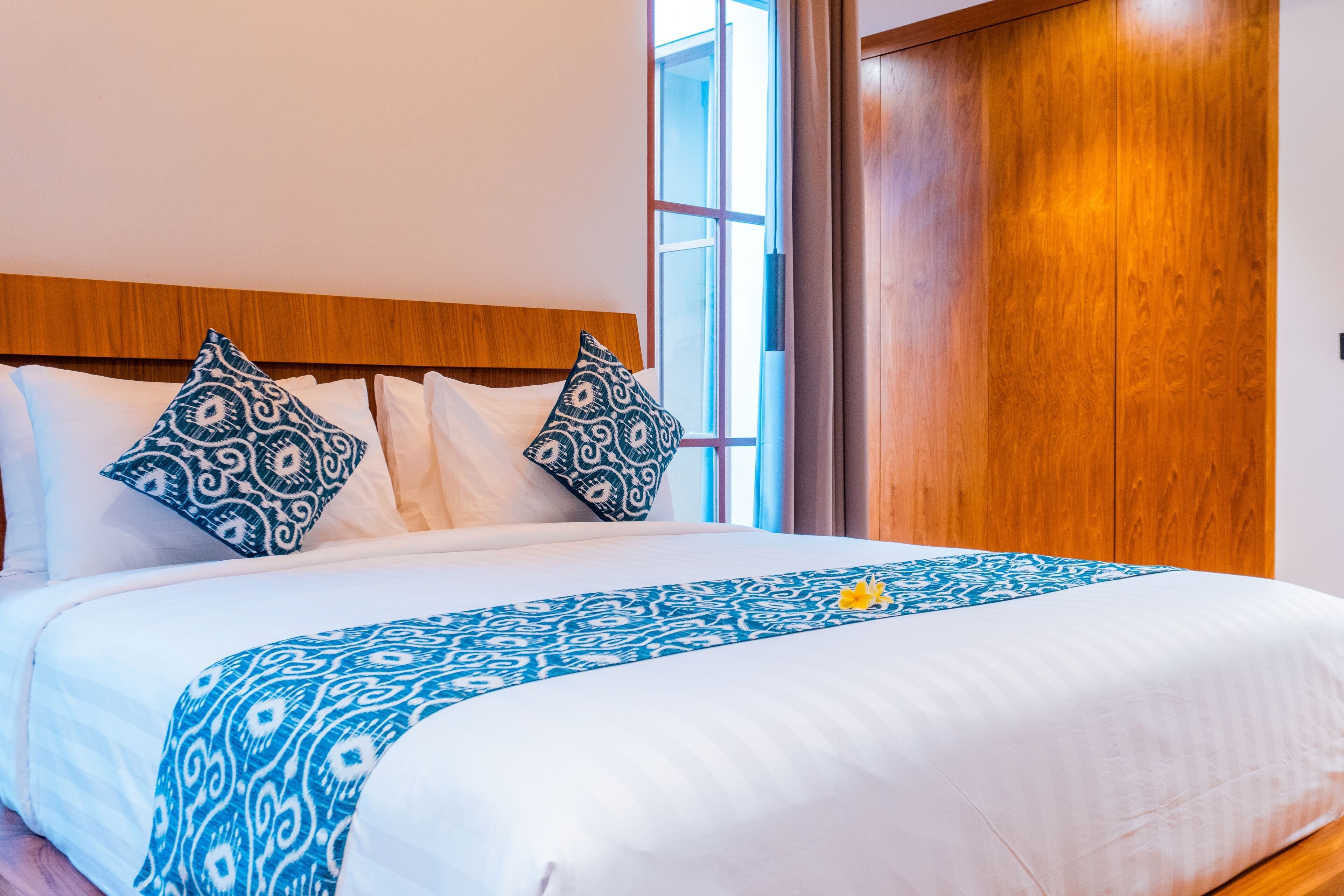 A neatly made bed with patterned pillows and a blue and white bedspread, a wooden headboard, and a window with curtains.