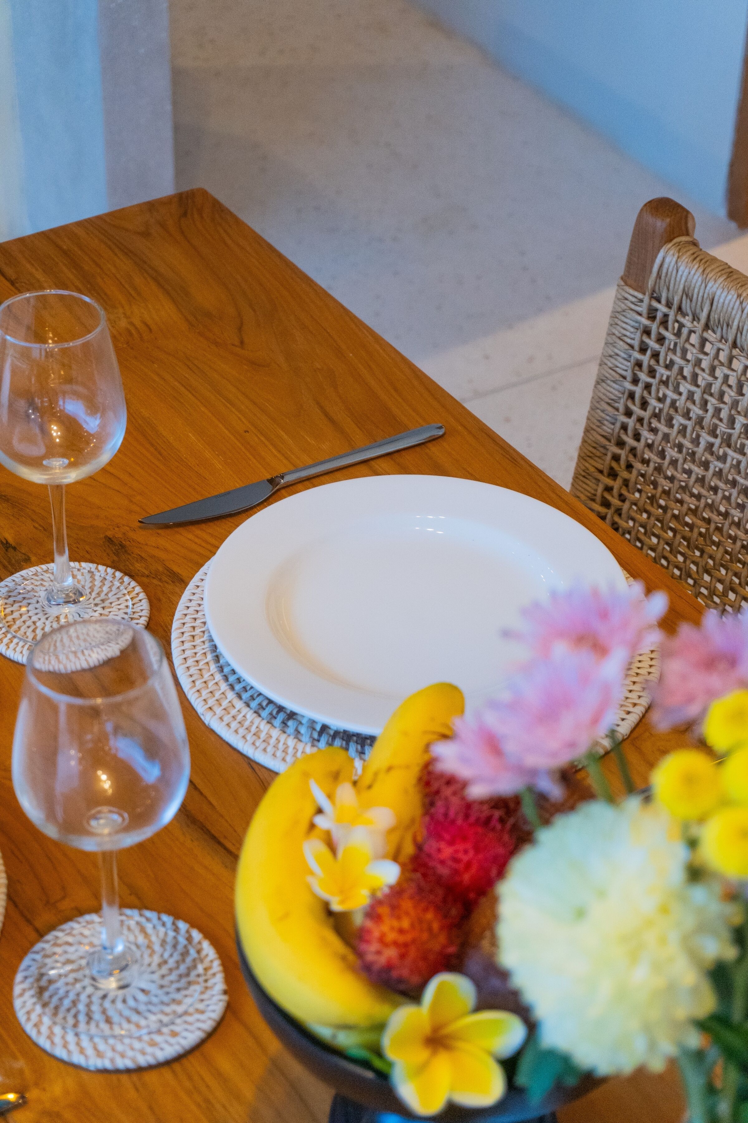 Ett träbord med en vit tallrik, två vinglas, en kniv och en skål med frukt och blommor.