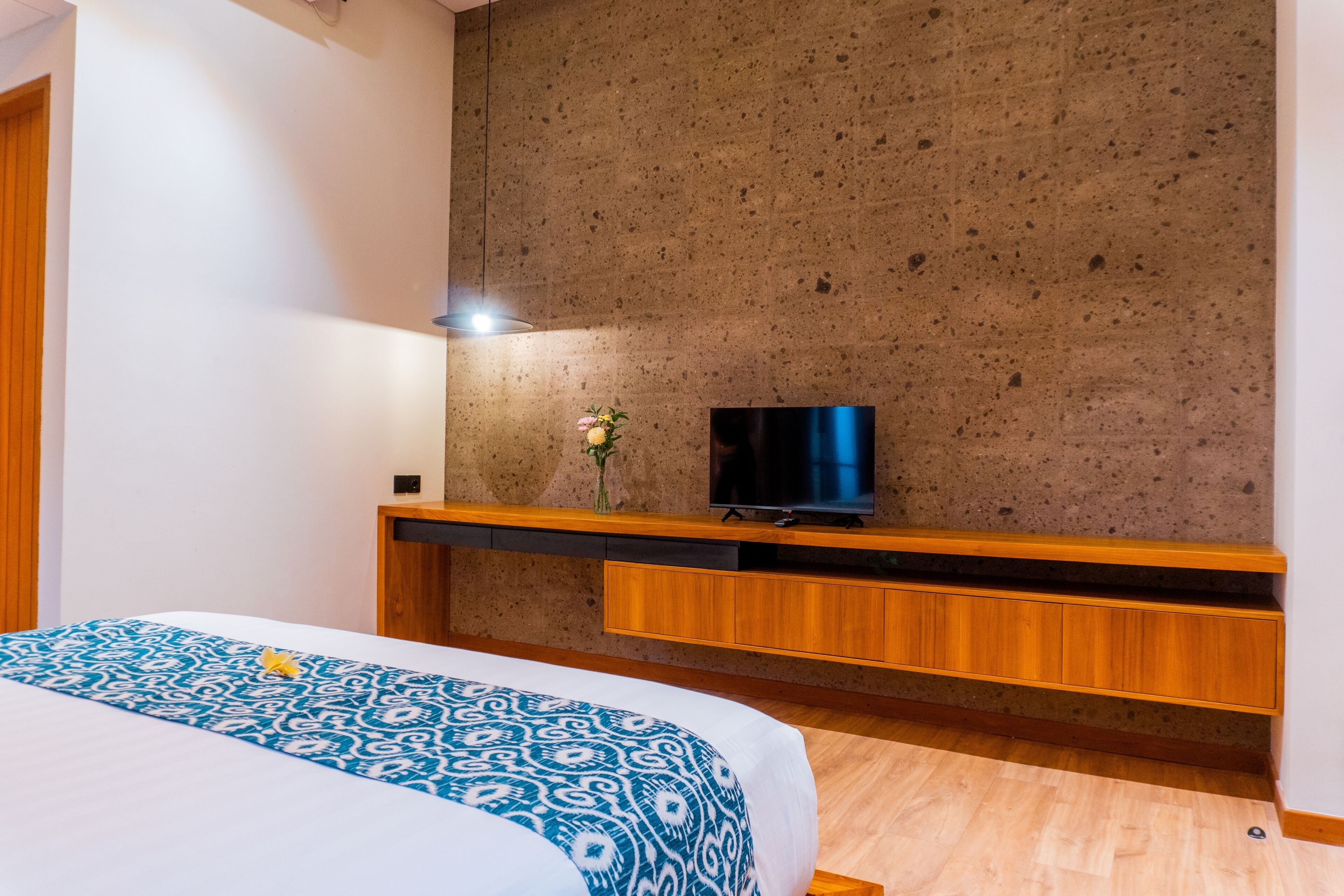 A hotel room with a bed, a flat-screen TV mounted on a textured wall, wooden furniture, and a small vase with flowers.
