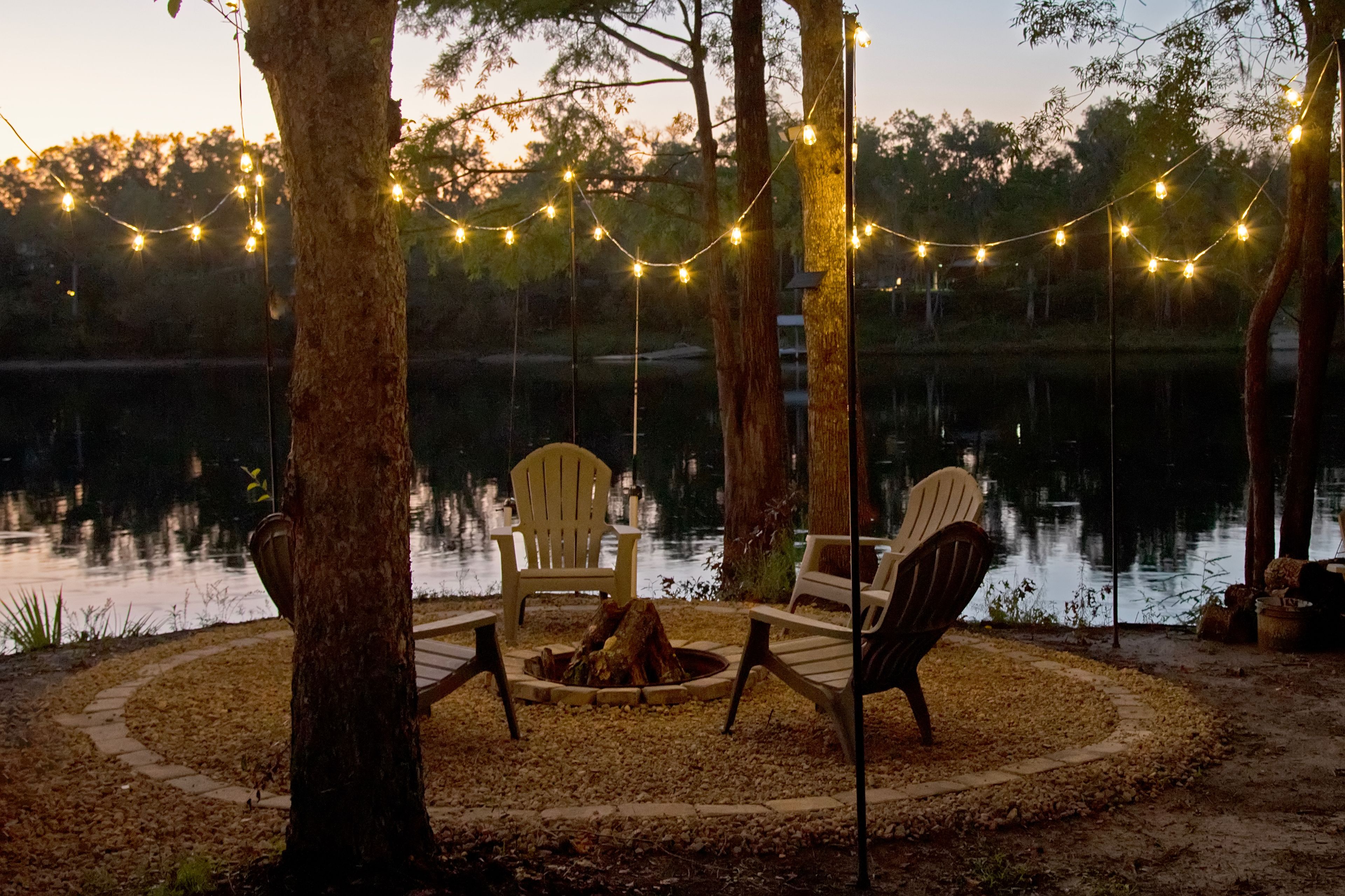 A lakeside seating area with two chairs and a fire pit, surrounded by trees and string lights.
