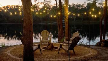 A lakeside seating area with two chairs and a fire pit, surrounded by trees and string lights.