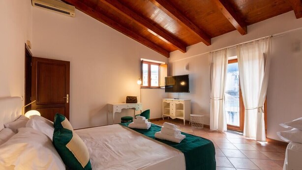 Een hotelkamer met een houten plafond, een bed met groen en witte beddengoed, een bureau met stoel, een televisie en een raam met gordijnen.